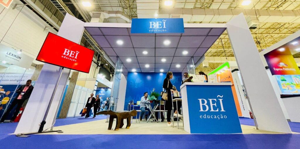 Foto mostra estande de feira com balcão azul escrito BEI em branco, um banco indígena e uma TV com tela vermelha escrito BEI em branco em primeiro plano