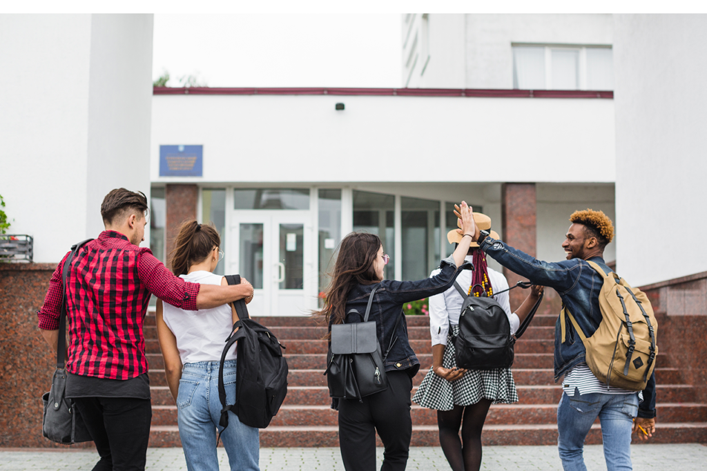 Cinco estudantes do Ensino Médio de costas caminham para a porta da escola. Dois estão abraçados e dois estão batendo as mãos no alto