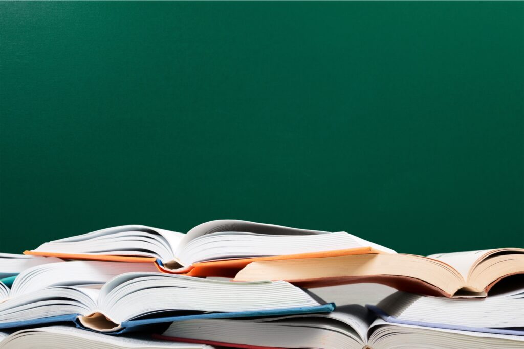 Foto mostra livros didáticos abertos em cima de uma mesa com uma lousa verde ao fundo
